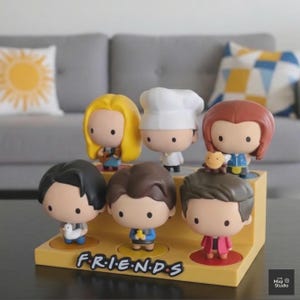 May include: A set of seven Friends character figurines displayed on a yellow stand. Each figurine has a unique hairstyle and outfit, with the Friends logo at the base. The figurines are in a cartoon style.