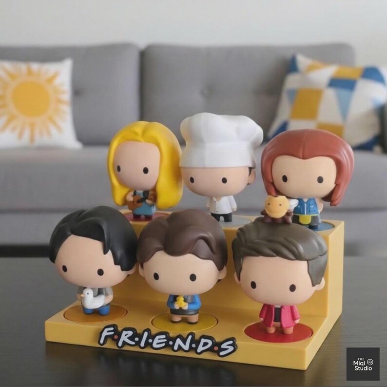 May include: A set of seven Friends character figurines displayed on a yellow stand. Each figurine has a unique hairstyle and outfit, with the Friends logo at the base. The figurines are in a cartoon style.