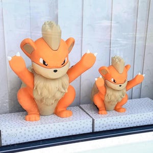 May include: Two orange and cream-colored Growlithe figurines in a crouching pose with arms raised. The larger figure is on the left and the smaller figure is on the right. Both have a tuft of fur on their heads and pointed ears with white claws.