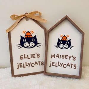 Puede incluir: Dos letreros rectangulares blancos con marcos marrones que muestran una ilustraci&oacute;n de un gato negro con un sombrero naranja. El letrero izquierdo tiene una cinta dorada y dice 'ELLIE'S JELLYCATS', el derecho 'MAISEY'S JELLYCATS' en letras marrones.
