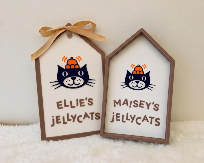 Puede incluir: Dos letreros rectangulares blancos con marcos marrones que muestran una ilustraci&oacute;n de un gato negro con un sombrero naranja. El letrero izquierdo tiene una cinta dorada y dice 'ELLIE'S JELLYCATS', el derecho 'MAISEY'S JELLYCATS' en letras marrones.