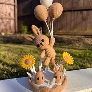 May include: A brown bunny figurine holding three balloons, with a basket base featuring two eggs, two bunnies, and two yellow sunflowers. The figurine is set on a white surface with a green grass background.