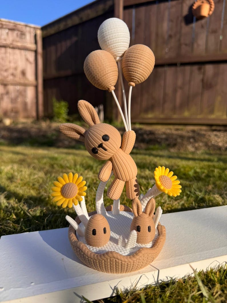 May include: A brown bunny figurine holding three balloons, with a basket base featuring two eggs, two bunnies, and two yellow sunflowers. The figurine is set on a white surface with a green grass background.