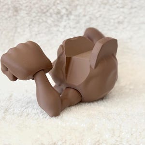 May include: A brown articulated doll body part, featuring a clenched fist and an arm. The figure is lying on its back, with the other arm and torso visible. The doll part is made of a solid, matte material.
