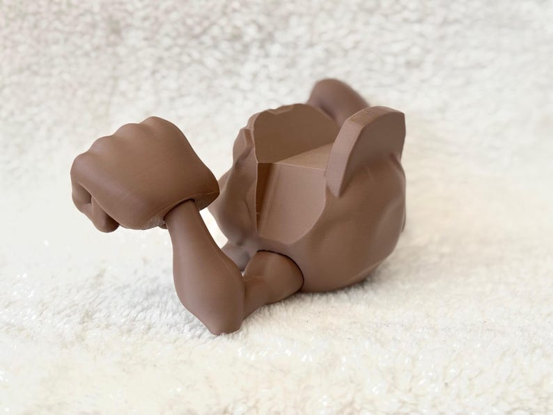 May include: A brown articulated doll body part, featuring a clenched fist and an arm. The figure is lying on its back, with the other arm and torso visible. The doll part is made of a solid, matte material.