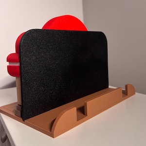 May include: A brown and black phone stand with a red accent. The stand has a rectangular black surface for holding a phone or tablet, supported by a brown base with a curved design. The stand is designed to hold a phone or tablet.