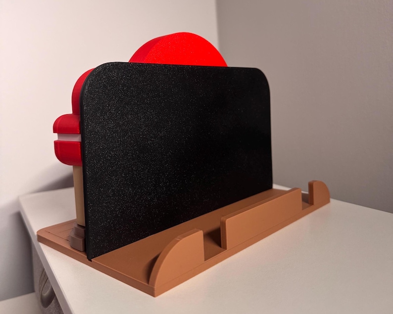 May include: A brown and black phone stand with a red accent. The stand has a rectangular black surface for holding a phone or tablet, supported by a brown base with a curved design. The stand is designed to hold a phone or tablet.