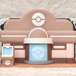 May include: A 3D-printed model of a building, featuring a tan and brown color scheme with white accents. The design incorporates a prominent circular logo, and a light blue screen. The model is designed to hold a gaming console.