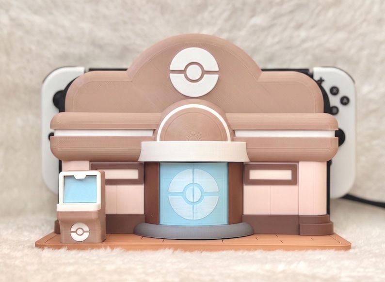 May include: A 3D-printed model of a building, featuring a tan and brown color scheme with white accents. The design incorporates a prominent circular logo, and a light blue screen. The model is designed to hold a gaming console.