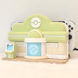 May include: A light green and beige gaming console dock shaped like a building with a central blue door, a white Pok&eacute; Ball emblem on the roof, and a small matching arcade cabinet accessory, holding a white handheld gaming console in the rear slot.