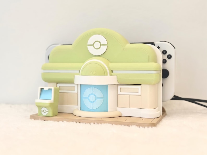 May include: A light green and beige gaming console dock shaped like a building with a central blue door, a white Pok&eacute; Ball emblem on the roof, and a small matching arcade cabinet accessory, holding a white handheld gaming console in the rear slot.