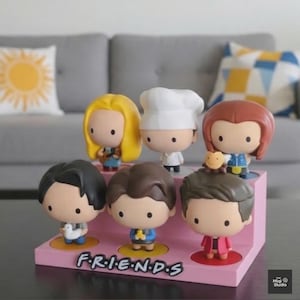 May include: A pink display stand featuring six cartoon figurines of characters from the TV show "Friends." The figurines have large heads and small bodies. The stand has the word "FRIENDS" in white letters.