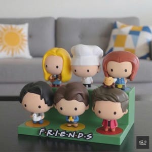 May include: A green display stand featuring six cartoon figurines of characters from the TV show "Friends." The figurines have large heads and small bodies. The word "FRIENDS" is printed on the stand.