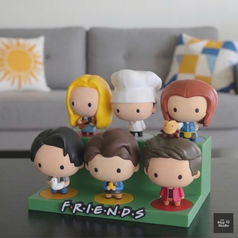 May include: A green display stand featuring six cartoon figurines of characters from the TV show "Friends." The figurines have large heads and small bodies. The word "FRIENDS" is printed on the stand.
