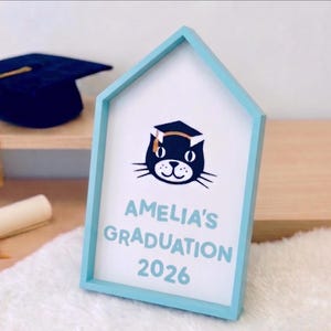 May include: A light blue framed sign with the text "AMELIA'S GRADUATION 2026" and a cat wearing a graduation cap. A navy blue graduation cap and a rolled diploma with a gold ribbon are in the background.