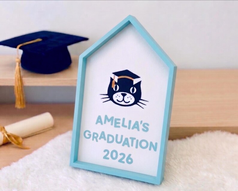 May include: A light blue framed sign with the text "AMELIA'S GRADUATION 2026" and a cat wearing a graduation cap. A navy blue graduation cap and a rolled diploma with a gold ribbon are in the background.