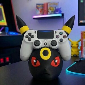Puede incluir: Un soporte negro para mando con tem&aacute;tica Umbreon con un mando de PlayStation blanco encima. El soporte tiene detalles amarillos y ojos rojos. Una figura de Pikachu es visible en el fondo.