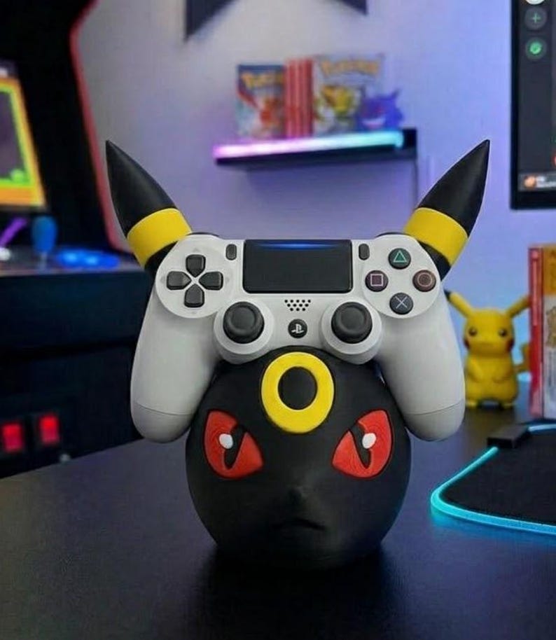 Puede incluir: Un soporte negro para mando con tem&aacute;tica Umbreon con un mando de PlayStation blanco encima. El soporte tiene detalles amarillos y ojos rojos. Una figura de Pikachu es visible en el fondo.