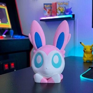 Puede incluir: Una figura de Sylveon blanca, rosa y azul. La figura tiene grandes orejas rosas con detalles azules y grandes ojos azules. Est&aacute; sobre una superficie negra, con una figura de Pikachu y cajas de videojuegos en el fondo.