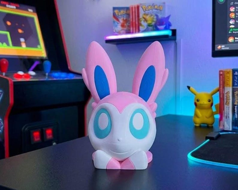 Puede incluir: Una figura de Sylveon blanca, rosa y azul. La figura tiene grandes orejas rosas con detalles azules y grandes ojos azules. Est&aacute; sobre una superficie negra, con una figura de Pikachu y cajas de videojuegos en el fondo.