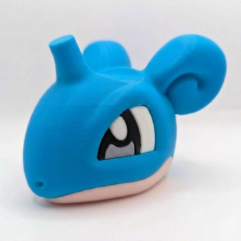 May include: A 3D-printed blue Lapras head figurine with a white, black, and gray eye, and a pink base. The figurine has a smooth texture and is designed with rounded features.