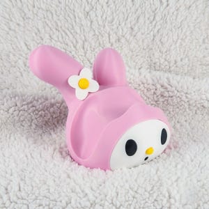 May include: A pink rabbit-shaped character figure with a white face, black oval eyes, and a small yellow nose. The figure features a white flower with a yellow center attached to one of its long, floppy ears, and a small black mouth detail.