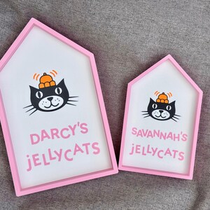 May include: Two house-shaped signs with pink frames and white interiors. Each sign features a black cat illustration wearing an orange hat. The larger sign reads "DARCY'S JELLYCATS" in pink, while the smaller one says "SAVANNAH'S JELLYCATS".