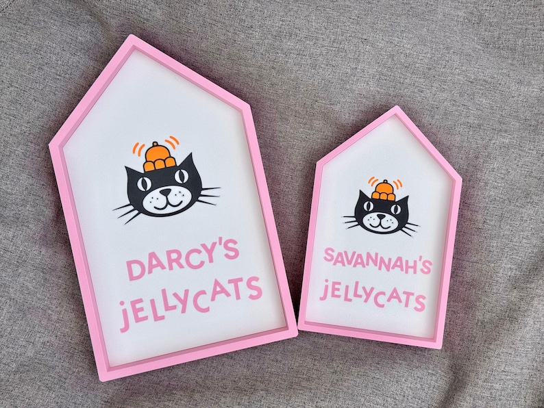 May include: Two house-shaped signs with pink frames and white interiors. Each sign features a black cat illustration wearing an orange hat. The larger sign reads "DARCY'S JELLYCATS" in pink, while the smaller one says "SAVANNAH'S JELLYCATS".