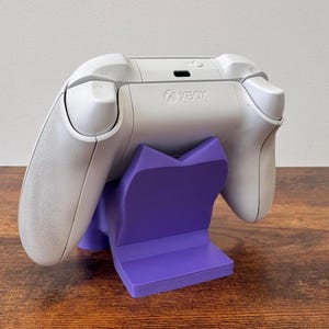 May include: A white Xbox wireless controller resting on a solid purple plastic display stand. The controller features the Xbox logo on the top center, with shoulder buttons and triggers visible. The stand has a V-shaped cradle design.