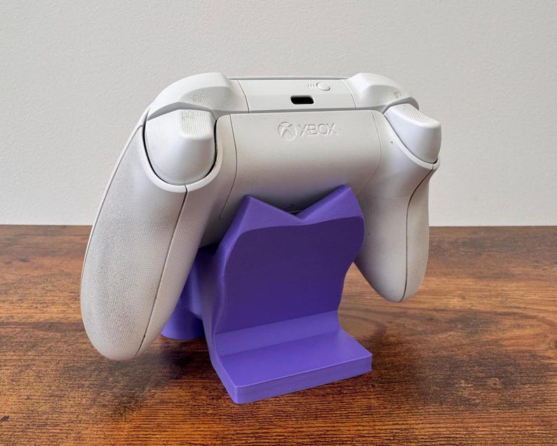 May include: A white Xbox wireless controller resting on a solid purple plastic display stand. The controller features the Xbox logo on the top center, with shoulder buttons and triggers visible. The stand has a V-shaped cradle design.