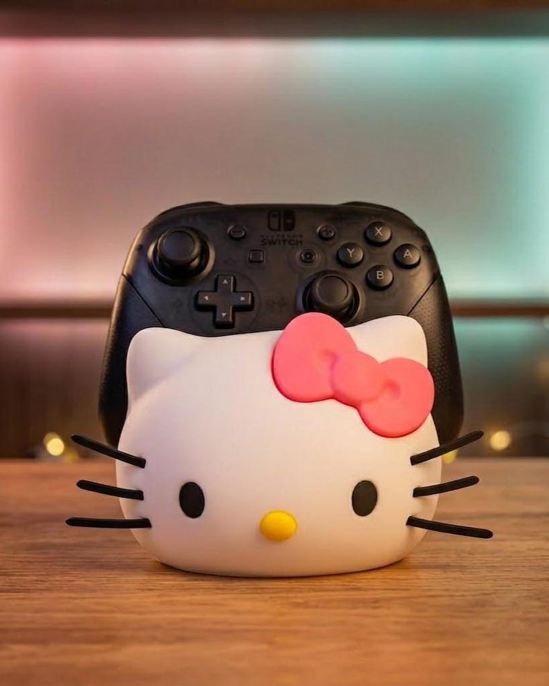 May include: A white Hello Kitty-themed Nintendo Switch controller holder with a pink bow, black whiskers, and black eyes. A black Nintendo Switch Pro Controller is behind the holder. The holder has a yellow nose and is on a wooden surface.
