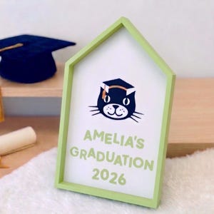 May include: A light green framed sign with the text "AMELIA'S GRADUATION 2026" and a cartoon cat wearing a graduation cap. A graduation cap and diploma are in the background.