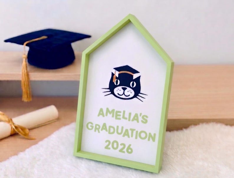 May include: A light green framed sign with the text "AMELIA'S GRADUATION 2026" and a cartoon cat wearing a graduation cap. A graduation cap and diploma are in the background.