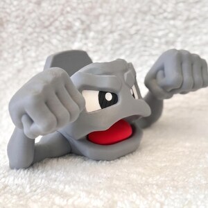 May include: A gray figurine of a cartoon character with a rocky appearance. The character has a wide open mouth with a red interior, white eyes, and clenched fists. The figurine is on a white, textured surface.