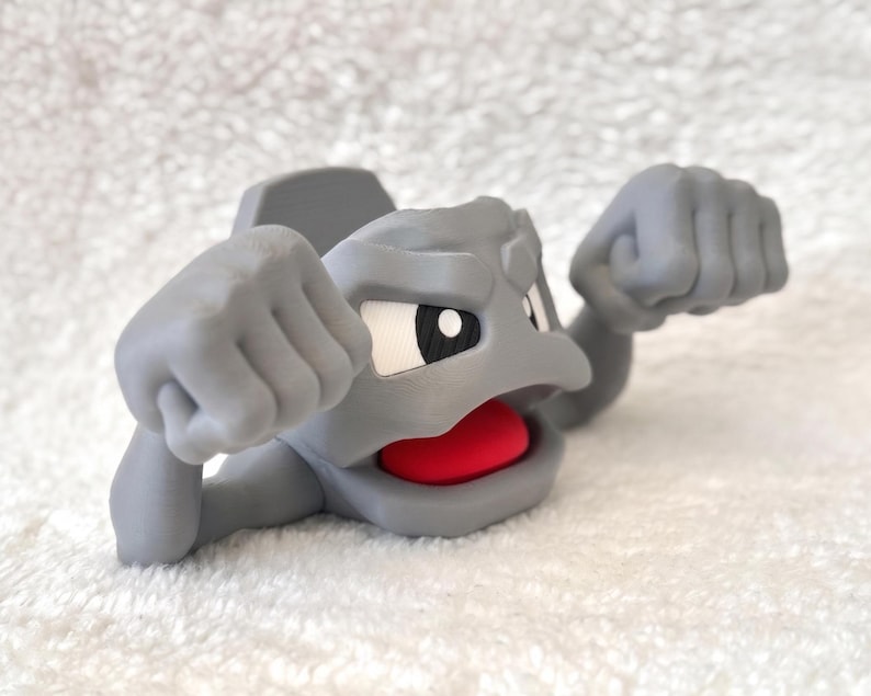 May include: A gray figurine of a cartoon character with a rocky appearance. The character has a wide open mouth with a red interior, white eyes, and clenched fists. The figurine is on a white, textured surface.