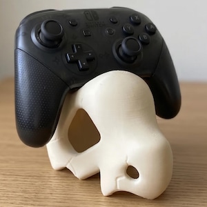 May include: A black Nintendo Switch Pro controller resting on a cream-colored, 3D-printed controller stand. The stand has a unique, abstract design with rounded shapes and a hole. The controller is black with visible buttons and joysticks.