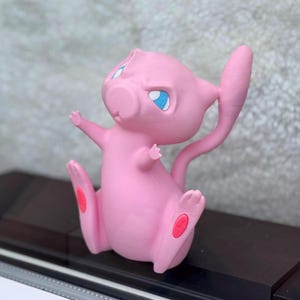 Puede incluir: Una figura rosa de Mew con grandes ojos azules, orejas puntiagudas y una cola larga y delgada. El personaje est&aacute; representado en posici&oacute;n sentada con los brazos levantados y peque&ntilde;as almohadillas ovaladas rosas visibles en las plantas de los pies.