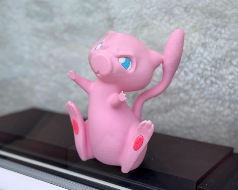 Puede incluir: Una figura rosa de Mew con grandes ojos azules, orejas puntiagudas y una cola larga y delgada. El personaje est&aacute; representado en posici&oacute;n sentada con los brazos levantados y peque&ntilde;as almohadillas ovaladas rosas visibles en las plantas de los pies.
