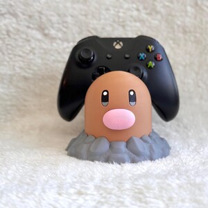 May include: A black gaming controller resting on a tan, cylindrical character stand with a pink nose and dark eyes, set upon a gray, textured base that resembles rocky ground. The controller features a central Xbox logo and colored buttons.