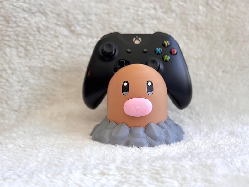 May include: A black gaming controller resting on a tan, cylindrical character stand with a pink nose and dark eyes, set upon a gray, textured base that resembles rocky ground. The controller features a central Xbox logo and colored buttons.