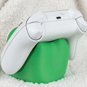 Puede incluir: Un mando de videojuegos inal&aacute;mbrico de color blanco apoyado sobre un soporte ergon&oacute;mico de color verde s&oacute;lido y forma redondeada. El mando cuenta con un puerto de carga USB-C y un peque&ntilde;o bot&oacute;n circular de sincronizaci&oacute;n en el borde superior.
