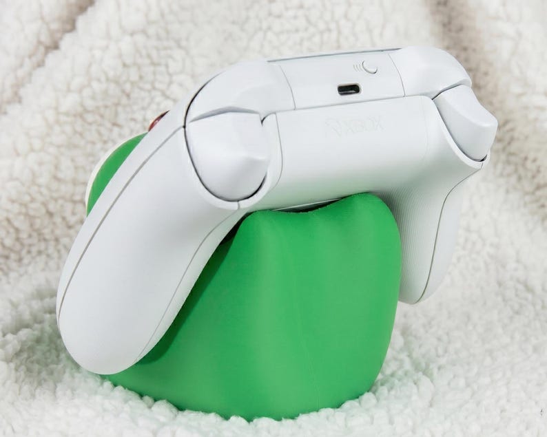 Puede incluir: Un mando de videojuegos inal&aacute;mbrico de color blanco apoyado sobre un soporte ergon&oacute;mico de color verde s&oacute;lido y forma redondeada. El mando cuenta con un puerto de carga USB-C y un peque&ntilde;o bot&oacute;n circular de sincronizaci&oacute;n en el borde superior.