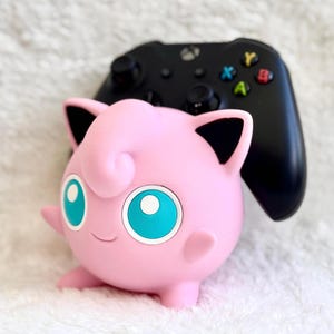 Peut inclure: Une figurine de personnage sph&eacute;rique rose avec de grands yeux bleu sarcelle, des oreilles pointues avec des d&eacute;tails int&eacute;rieurs noirs et un petit sourire, plac&eacute;e devant une manette de jeu vid&eacute;o sans fil noire avec des boutons color&eacute;s.