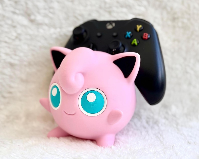 Peut inclure: Une figurine de personnage sph&eacute;rique rose avec de grands yeux bleu sarcelle, des oreilles pointues avec des d&eacute;tails int&eacute;rieurs noirs et un petit sourire, plac&eacute;e devant une manette de jeu vid&eacute;o sans fil noire avec des boutons color&eacute;s.