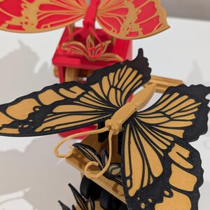 May include: Close-up of a decorative butterfly mobile. The butterflies have intricate wing designs with black outlines and gold and red coloring. The mobile is made of wood and has a detailed, layered design.