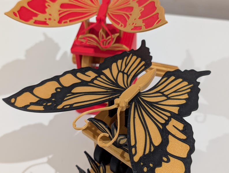 May include: Close-up of a decorative butterfly mobile. The butterflies have intricate wing designs with black outlines and gold and red coloring. The mobile is made of wood and has a detailed, layered design.