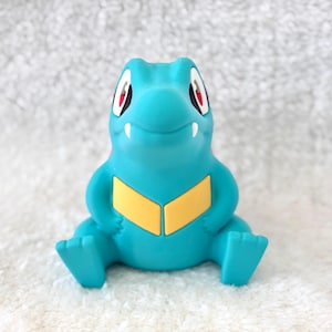 May include: A turquoise figurine of a cartoon crocodile-like creature with red eyes and a yellow chest. The figurine is sitting with its legs outstretched and arms at its sides. The background is a white, textured surface.