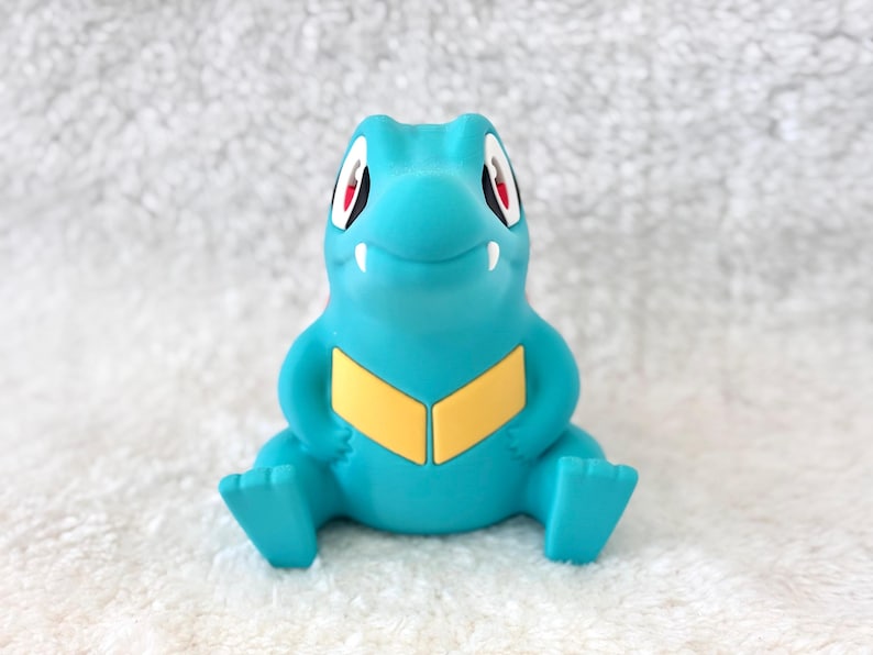 May include: A turquoise figurine of a cartoon crocodile-like creature with red eyes and a yellow chest. The figurine is sitting with its legs outstretched and arms at its sides. The background is a white, textured surface.