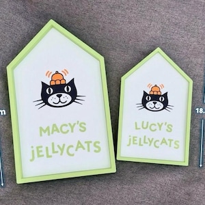 May include: Two house-shaped signs with a light green border. The signs feature a black cat illustration wearing a hat and the text "MACY'S JELLYCATS" and "LUCY'S JELLYCATS". The larger sign is 10.3 inches tall, and the smaller sign is 7.4 inches tall.
