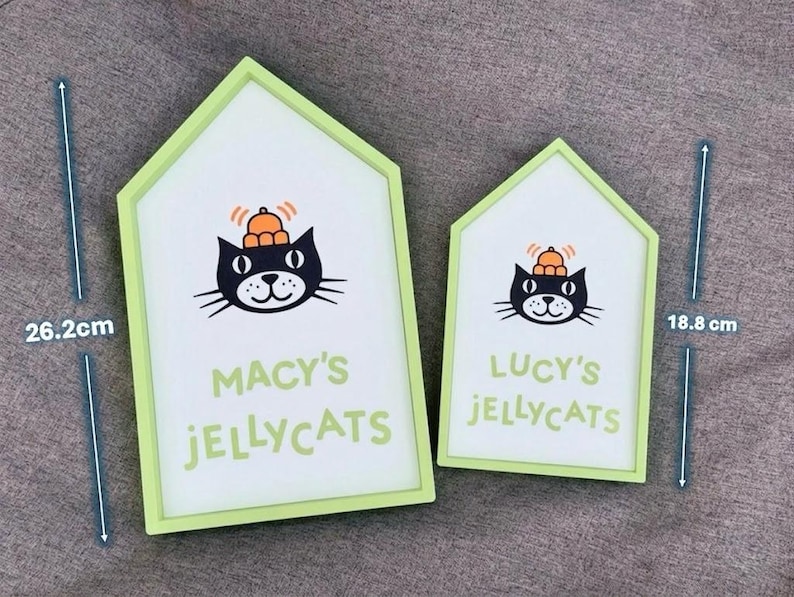 May include: Two house-shaped signs with a light green border. The signs feature a black cat illustration wearing a hat and the text "MACY'S JELLYCATS" and "LUCY'S JELLYCATS". The larger sign is 10.3 inches tall, and the smaller sign is 7.4 inches tall.